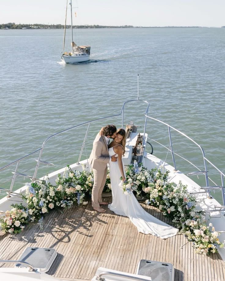 Bride and groom embracing on a yacht deck decorated with elegant floral arrangements, with the ocean and sailboat in the background