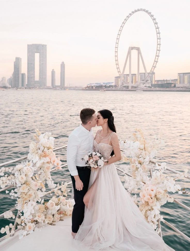 Bride and groom kissing on a yacht decorated with white and blush flowers, with Dubai skyline and Ain Dubai Ferris wheel in the background at sunset.
