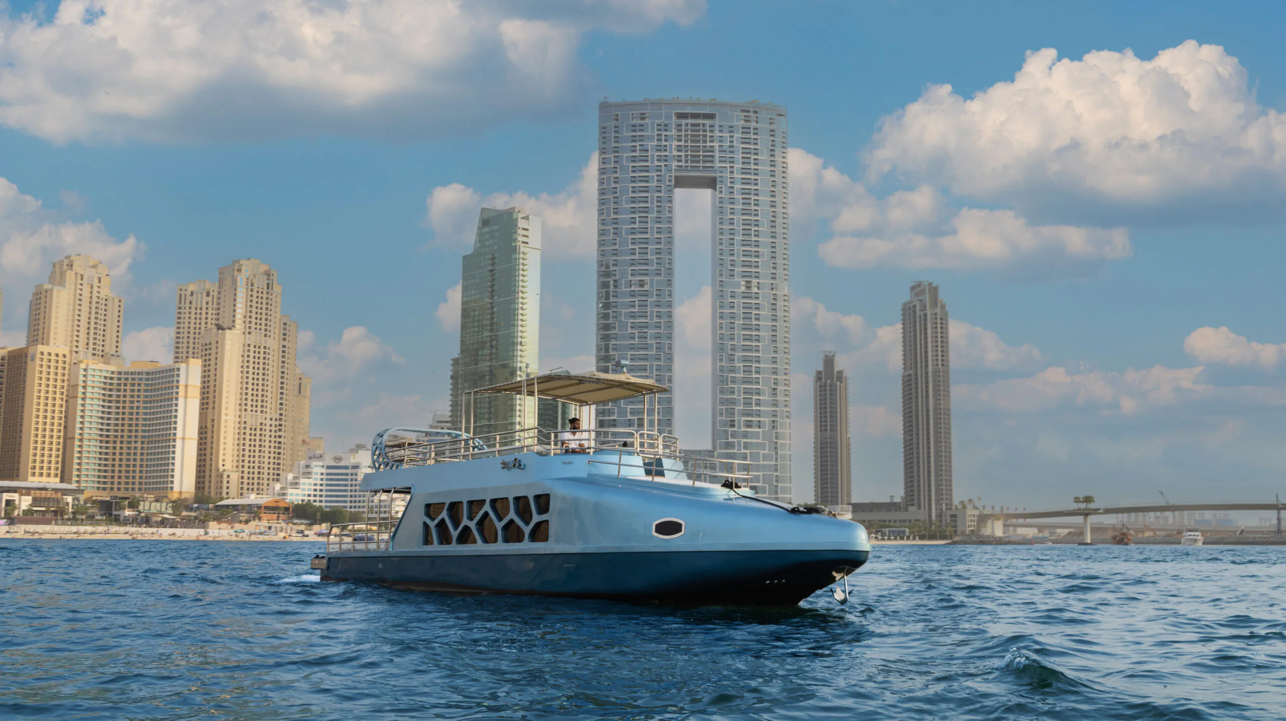 A modern luxury yacht cruising on blue water with Dubai Marina skyscrapers and waterfront skyline in the background under a clear sky.