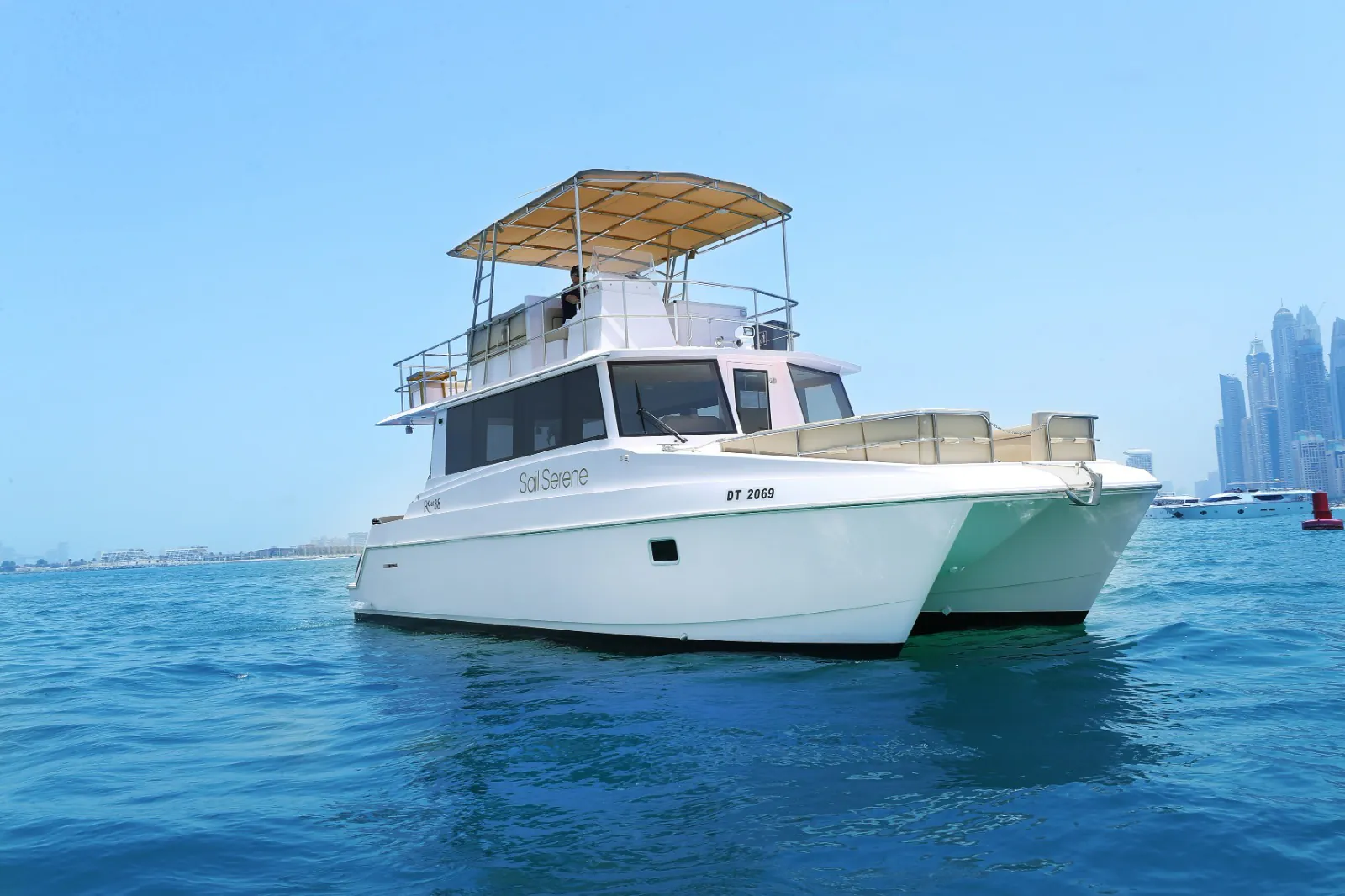 A white catamaran yacht floating on calm blue water, featuring a shaded upper deck and the Dubai skyline visible in the distance.
