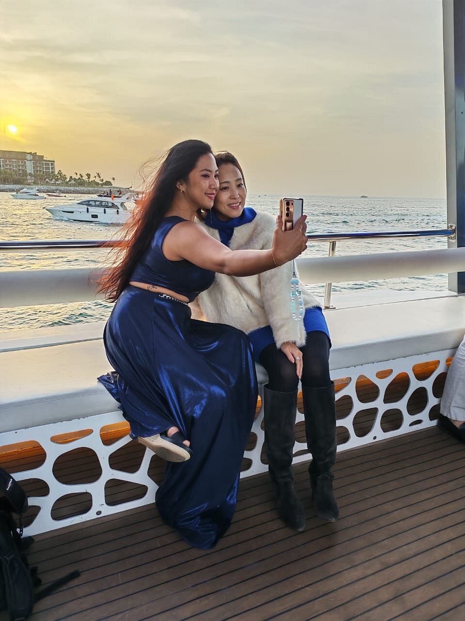 Two guests photographing themselves on the yacht deck with the sunset and Dubai coastline behind them